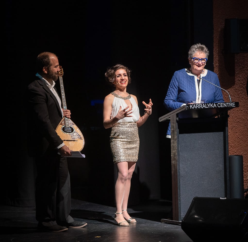 A man with an ornate guitar, stands next to a performer in a golden dress, they laugh with Showcase Victoria MC Christine Dunstan, who is standing behind a lecturn in a blue coat and matching blue glasses.